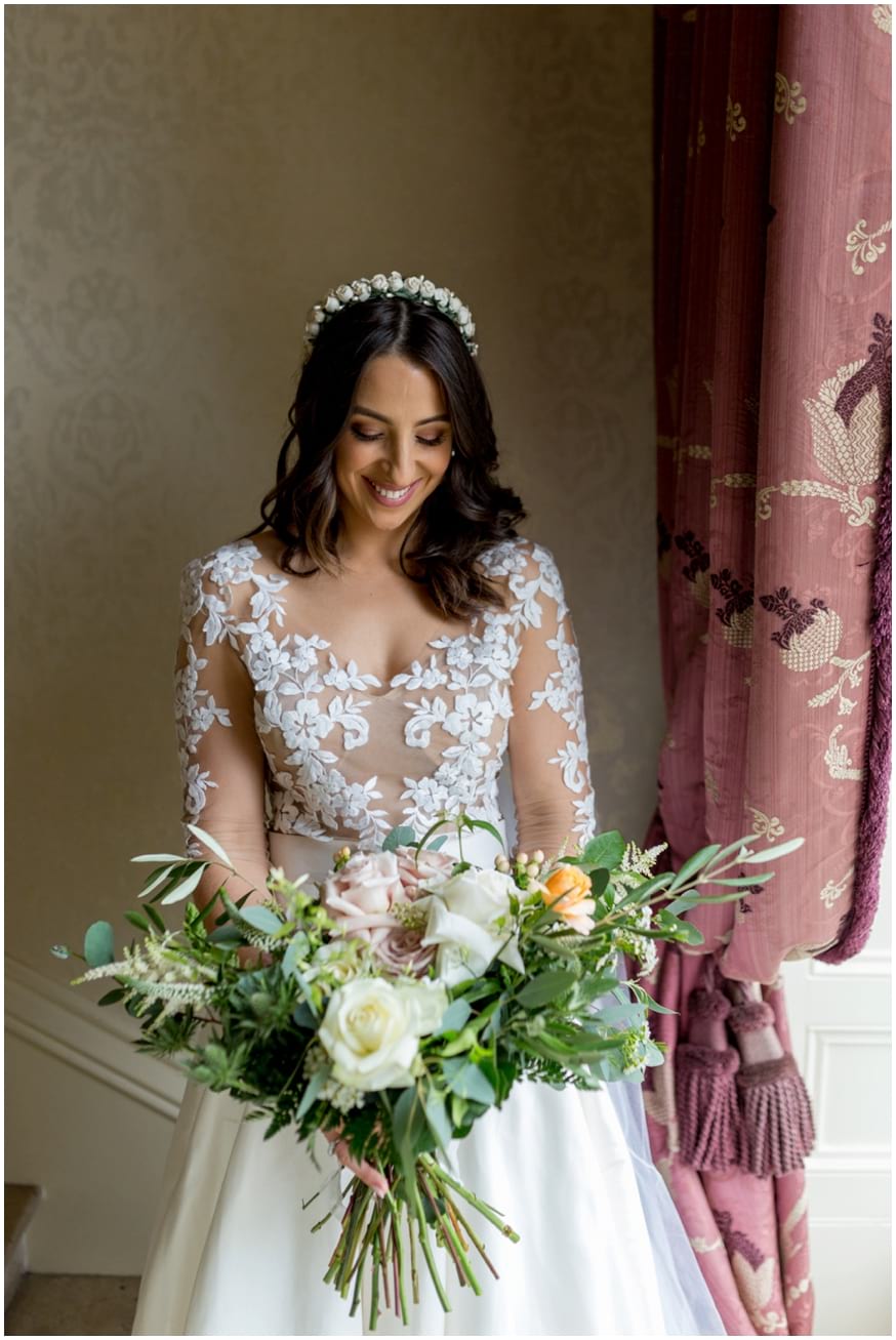 Bride stands by a window with her light pastel coloured bouquet