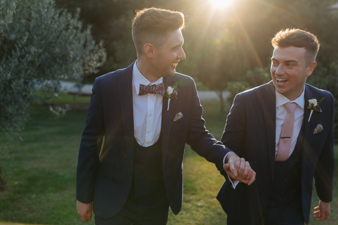 Same sex grooms hold hands as they walk through the beautiful gardens at Borgo Di Tragliata, Rome