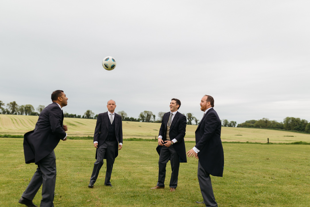 Groom and Groomsmen play football in their three piece wedding suits.